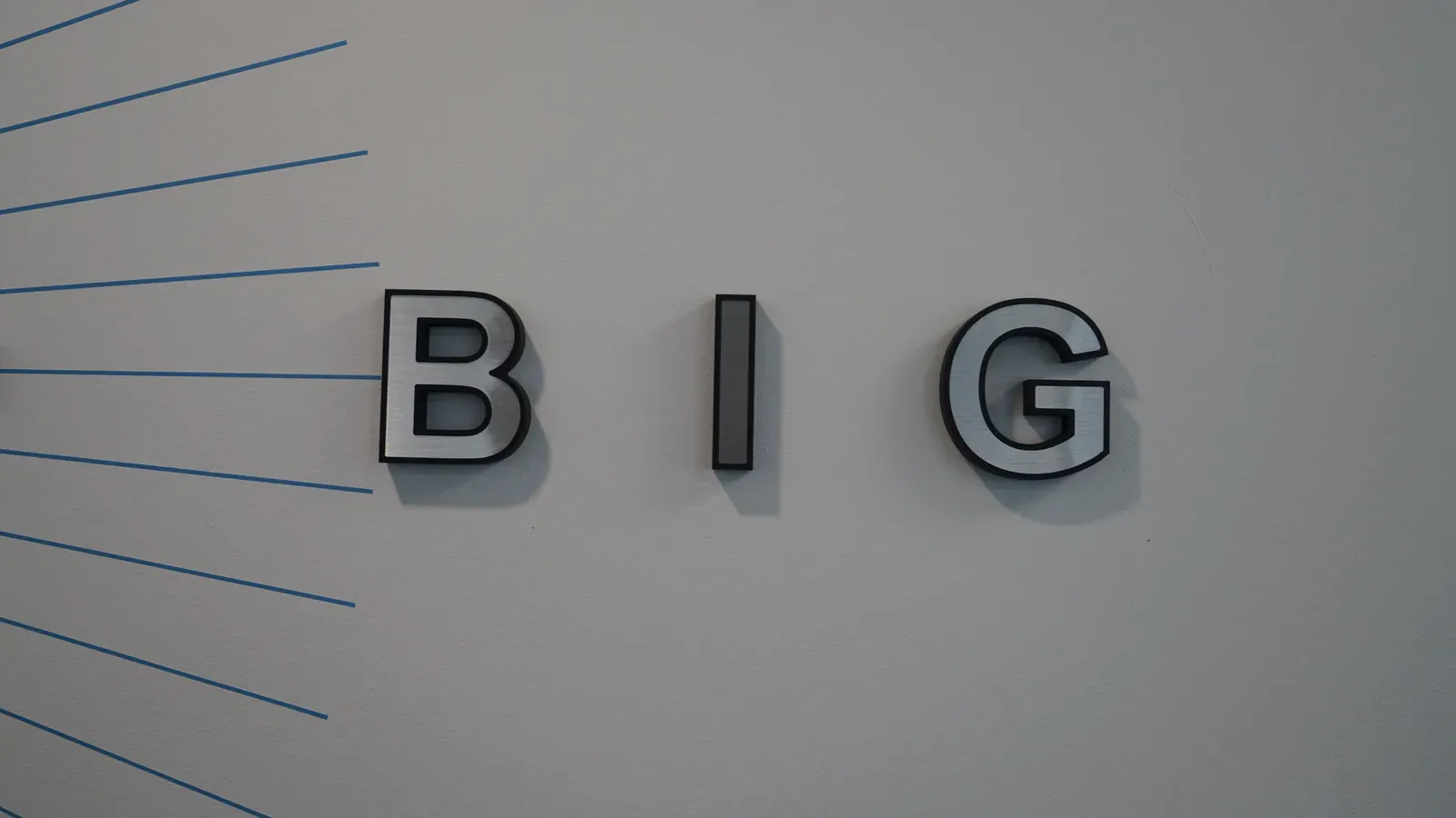 Mot BIG en lettres argentées en relief sur un mur gris traversé de lignes