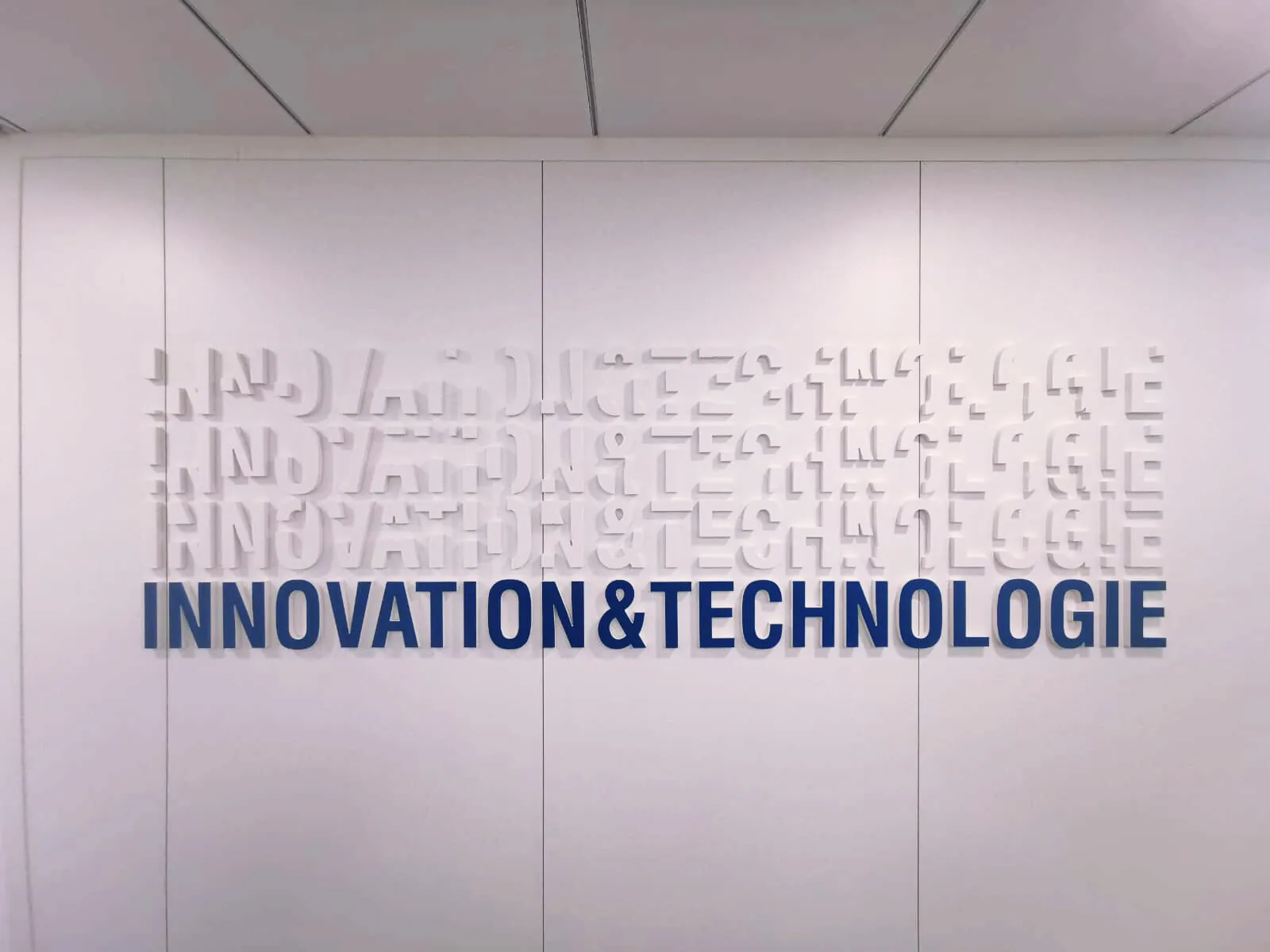 Mots Innovation & Technologie en lettres en relief, blanches en écho et bleues au premier plan, sur un mur blanc