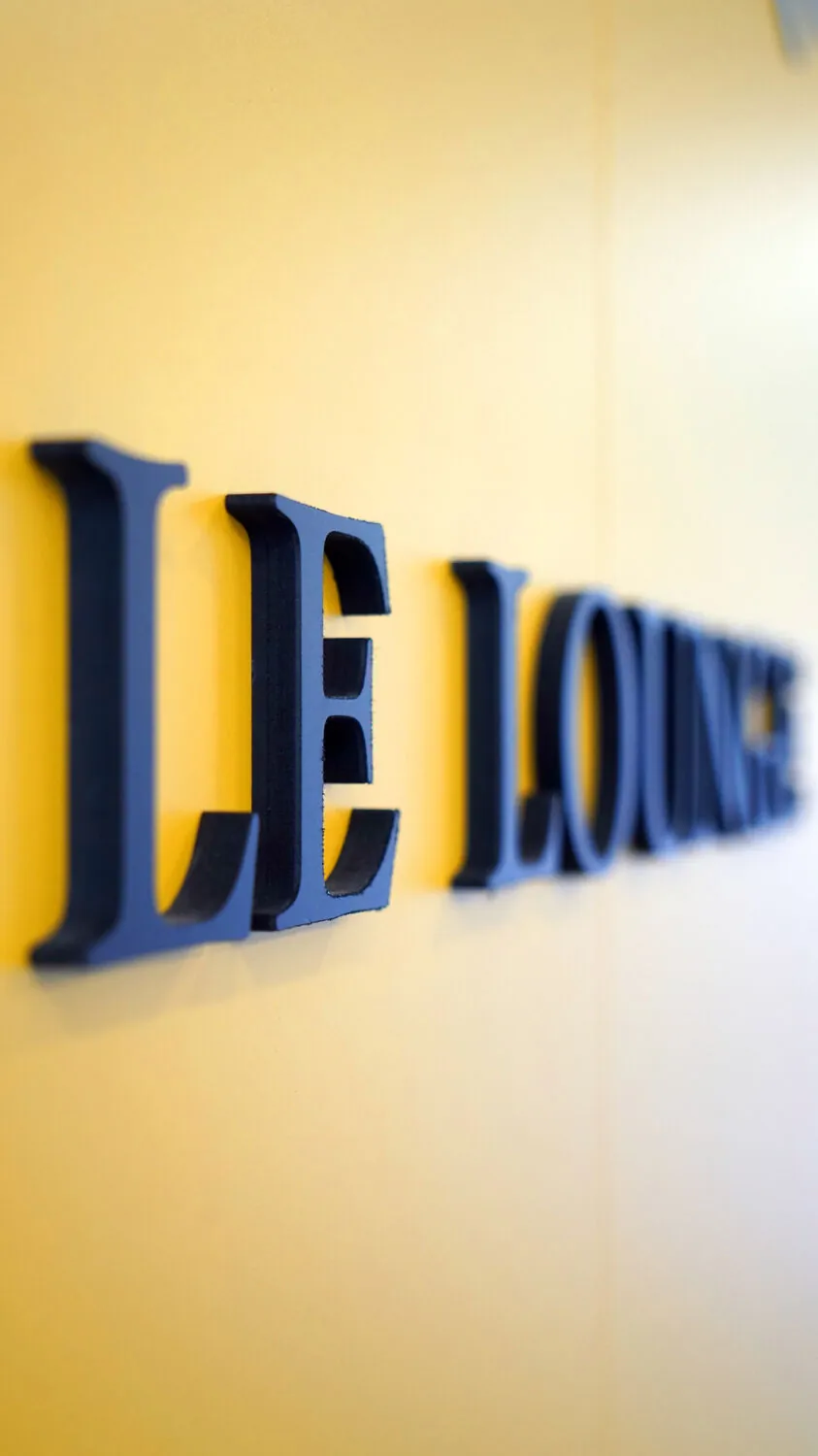 Lettres bleues LE LOUNGE en PVC, vues de profil sur un mur jaune