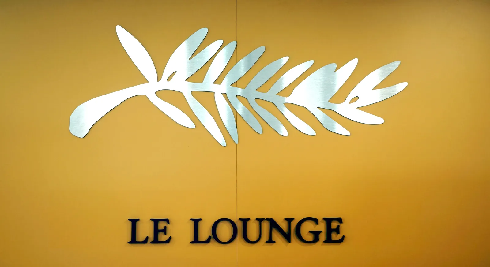 Branche d'olivier en Dibond effet miroir et mot LE LOUNGE en lettres noires sur un mur jaune