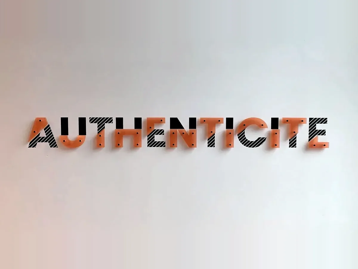 Mot AUTHENTICITÉ en lettres plexiglass orange et adhésif rayé noir sur une cloison blanche