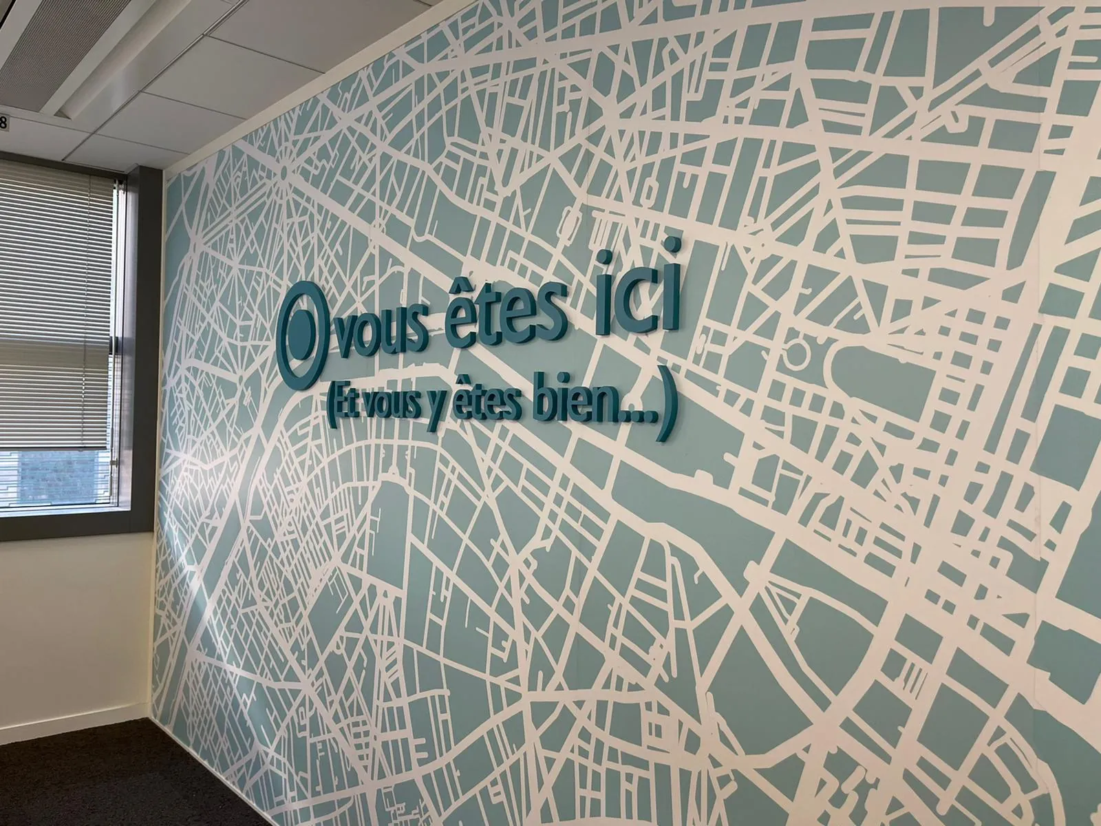 Texte « Vous êtes ici (et vous y êtes bien) » en lettres bleu-vert en relief sur une fresque de plan de ville
